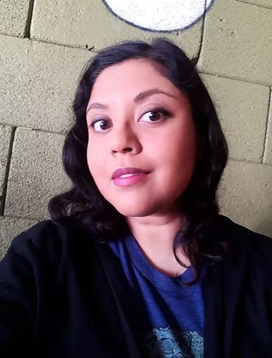 Premee Mohamed is an Indo-Caribbean femme with large, dark eyes, wavy shoulder-length hair, and tan skin. In this image, she is wearing a navy blue cardigan, a deep blue t-shirt, and an enigmatic smile. 