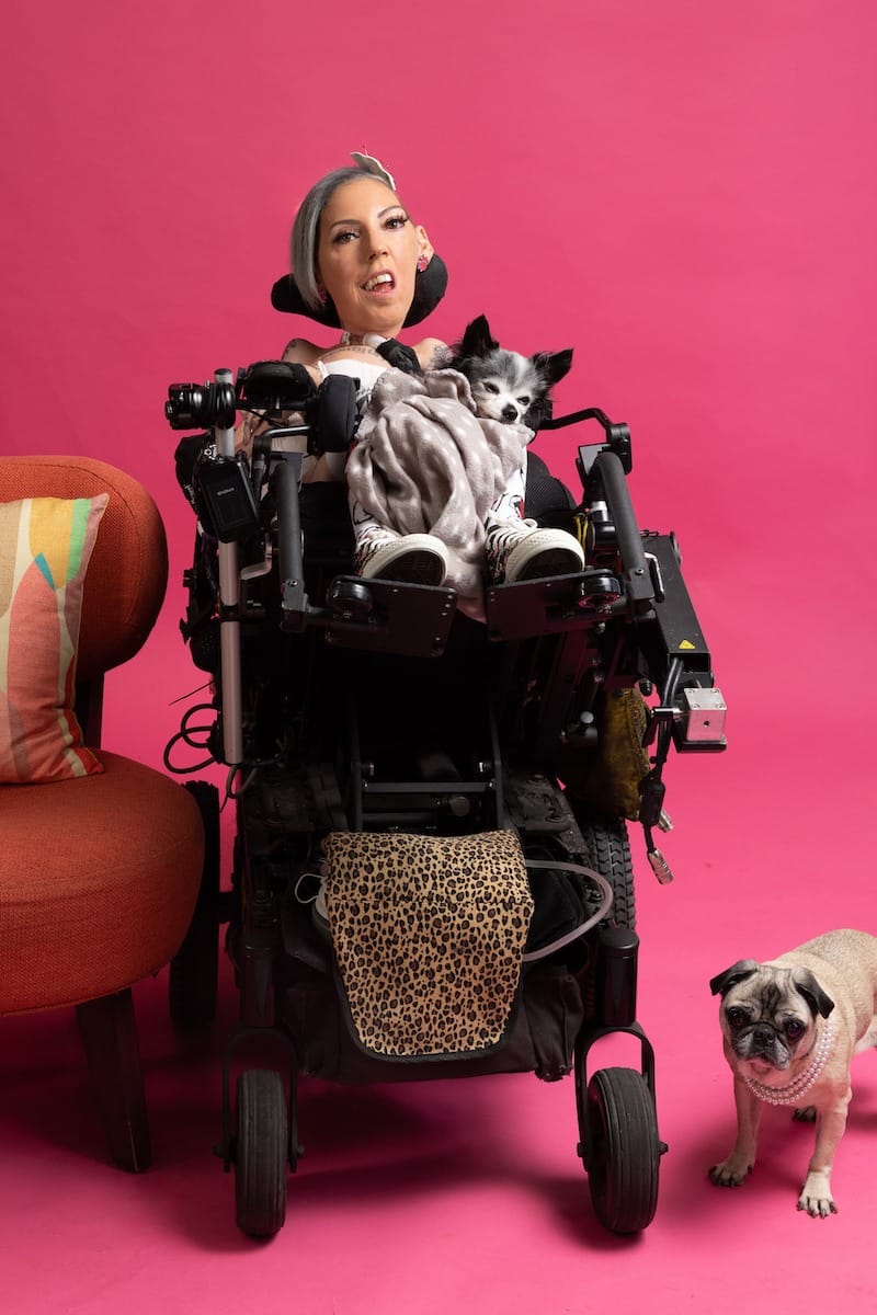 Annie Wilson in her wheelchair and in her lap is a small sleepy dog wrapped in a fuzzy grey blanket. On the floor, near the wheelchair, is a pug dog wearing a pearl necklace.