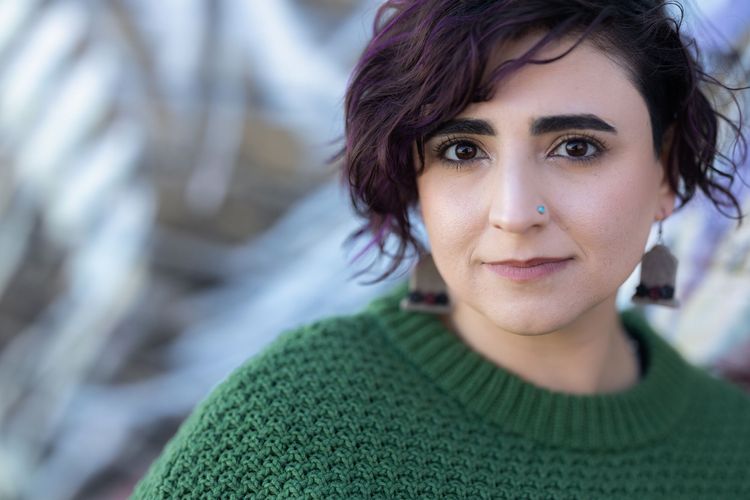 Naseem Jamnia, a Persian person in a green knit sweater with a nose piercing, wavy hair, and tombstone-shaped earrings.