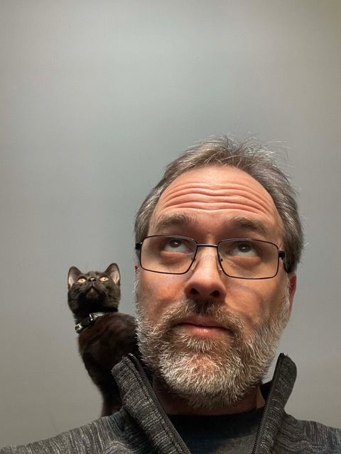 William Alexander, a man with short grey hair, a trim grey beard, and glasses. A black kitten looms over his shoulder. 
