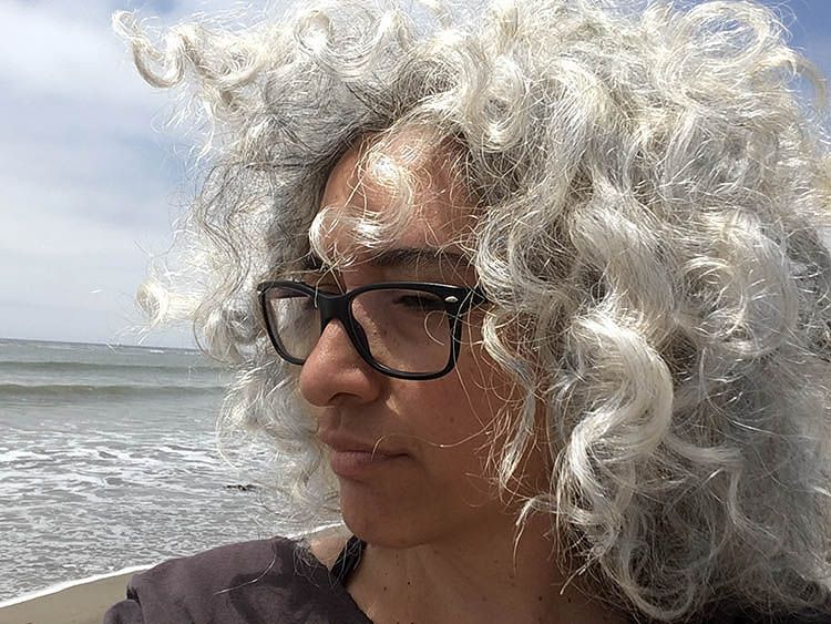 Profile view of author wearing glasses with the Pacific Ocean in the background. Her hair is wild. Sea-foam white curls.