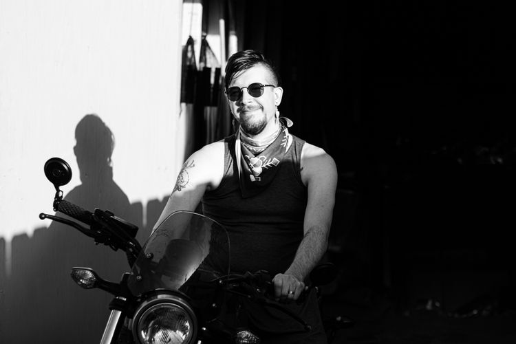 A smiling person with dark glasses, a goatee, and an undercut wearing a sleeveless top and sitting astride a motorcycle.