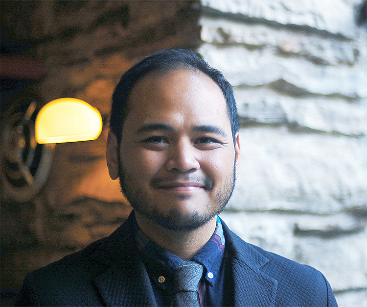 A smiling Filipino-American person with tan skin, short black hair, and very trim facial hair.
