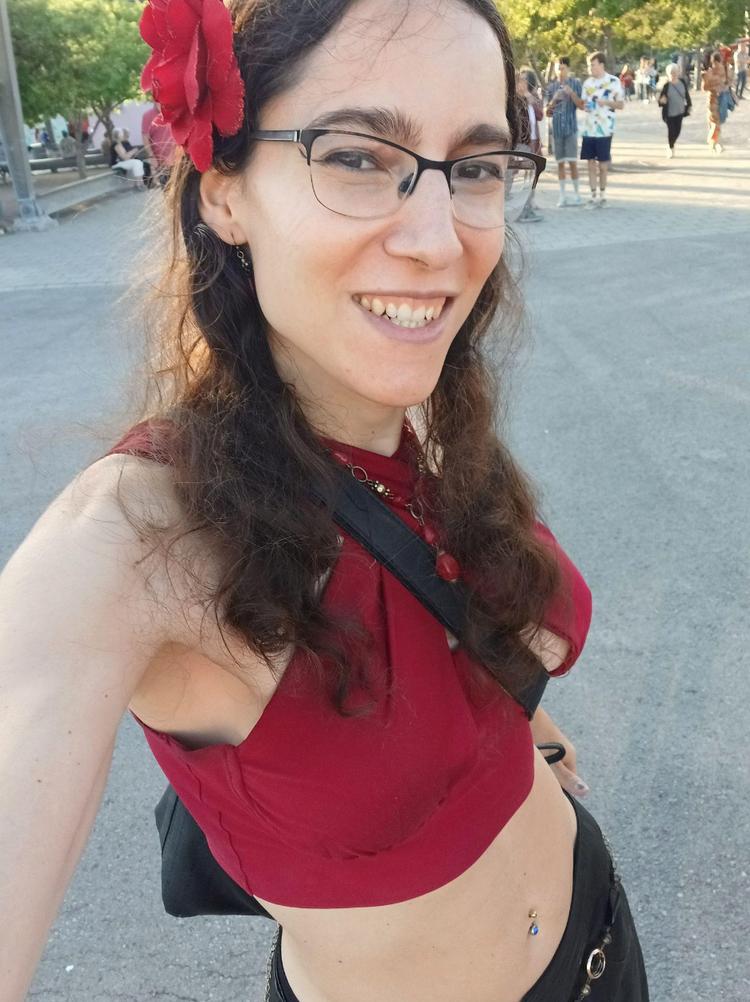 A smiling woman in a red crop top and black pants. She wears glasses and has a red flower in her long, wavy hair.