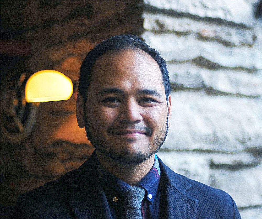 A smiling Filipino-American person with tan skin, short black hair, and very trim facial hair.